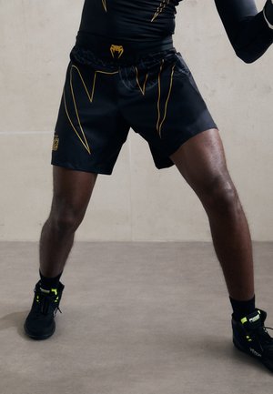 Athlete wearing black and yellow sports shorts and black Venum sneakers stands with legs apart on gray floor.