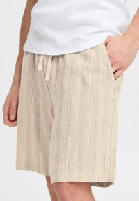 Shorts beige clair à rayures verticales, fabriqués en tissu texturé. Dotés d'une taille élastique avec cordon de serrage et de poches latérales.