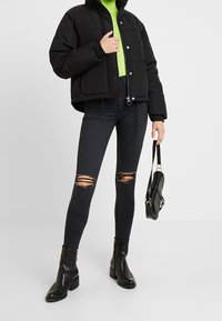 Černá péřová bunda s zkráceným designem, spojena s černými roztrhanými skinny džíny. Černé kotníkové boty a texturovaná černá kabelka dotvářejí celkový vzhled.