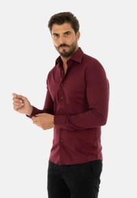 Camicia bordeaux a maniche lunghe realizzata in tessuto liscio, con colletto appuntito, bottoni sul davanti e polsini aderenti. Pantaloni neri indossati sotto.
