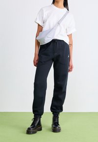 Schwarze Jogginghose mit elastischen Bündchen, weißem Logo, kombiniert mit einem weißen T-Shirt und silberner Umhängetasche. Schwarze Schnürstiefel vollenden den Look.