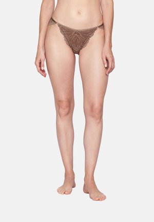 Woman wearing brown lace thong underwear, standing barefoot against a plain light background.