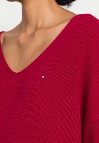 V-hals, losse gebreide trui in felrood, met een gemêleerd textuur en een klein logo-patch met blauwe, witte en rode accenten.