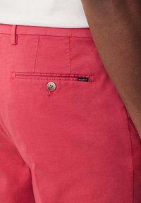 Rote Baumwollhosen mit taillierter Passform, ausgestattet mit einer Gesäßtasche mit Knopf und einem kleinen Markenlabel mit der Aufschrift "HACKETT" in der Nähe der Tasche.