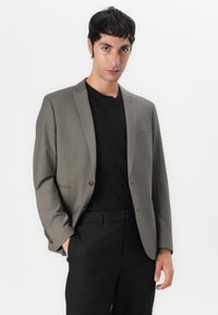 Blazer gris de tejido suave, con diseño de un solo botón, solapas con muesca y un bolsillo en el pecho, llevado sobre una camisa negra.