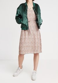 Veste bomber en satin vert portée sur une robe à motifs floraux avec des manches courtes. Des baskets blanches complètent la tenue. Bijoux simples visibles.