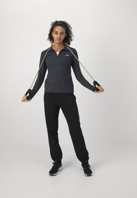 Μακρυμάνικο, σκούρο γκρι μπλουζάκι γυμναστικής με φερμουάρ και λογότυπο, συνδυασμένο με μαύρο παντελόνι φόρμας και μαύρα αθλητικά παπούτσια, κρατώντας λάστιχα αντίστασης.