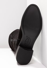 Botte en daim noire avec semelle lisse et fermeture éclair sur le côté, photographiée de la semelle tournée vers le haut sur fond blanc.