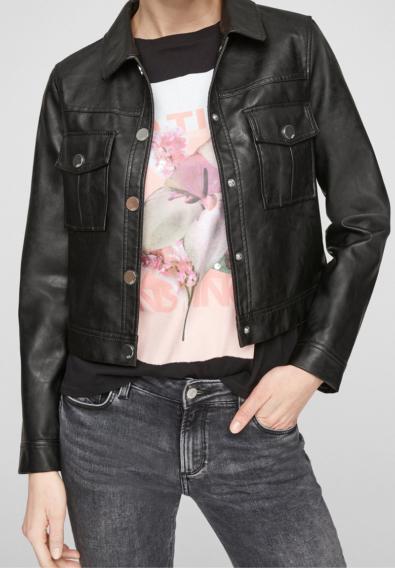 Chaqueta de cuero negra corta con dos bolsillos frontales, botones a presión plateados y una textura suave, llevada sobre una camiseta gráfica y jeans.