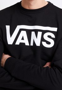 Persona con le braccia incrociate, che indossa una felpa nera con un grande logo bianco "VANS" sul petto e una sottile collana d'argento.