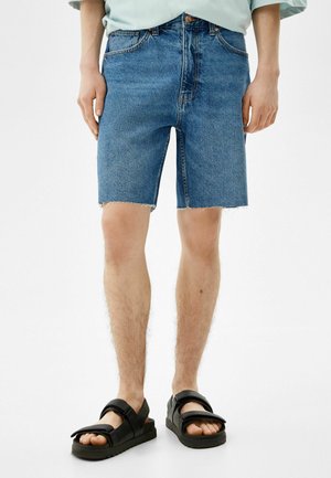 Denim-Shorts in mittlerem Waschung mit rohem Saum, kombiniert mit schwarzen Sandalen mit dicken Riemen und einer strukturierten Sohle.