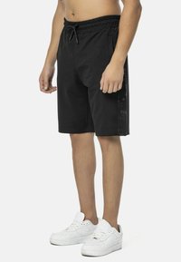 Schwarze Shorts mit elastischem Bund, Kordelzugverschluss und seitlichen Akzenten mit tonalem Textmuster. Das Material wirkt leicht und lässig.