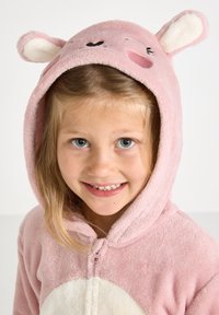 Una giovane ragazza sorride, indossando una tuta animalier rosa pallido con cappuccio, dotata di orecchie e dettagli facciali ricamati.