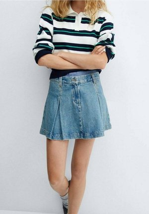 Osoba złożonymi ramionami, ubrana w pasiastą koszulę z długim rękawem, niebieską spódnicę dżinsową i szare skarpetki, stoi na tle jednolitego tła.