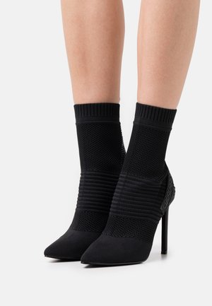 Svarta högklackade ankelskor med mesh och ribbat stickat mönster, spetsig tå och texturerade detaljer. Stiletto-klack och elegant profil.