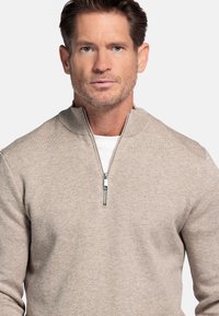 Lichtbeige ritstrui met een hoge kraag, voorzien van een gladde textuur en subtiel patroon, gecombineerd met een wit onderhemd.
