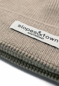 Dzianinowa czapka w jasnym beżu z teksturowanym wzorem. Posiada prostokątną metkę z napisem „slopes&town” oraz „AMSTERDAM” w czarnym tekście.
