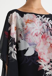 Svart transparent tygblus med stora blommönster i rosa och vita nyanser, med en avslappnad design och bred halsringning för enkel användning.