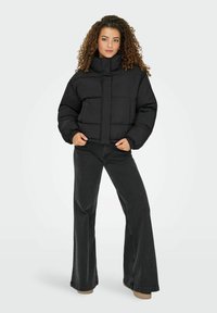 Kvinde med krøllet hår iført en sort dunjakke og mørke jeans med vide ben, stående med hænderne i lommerne på en lys baggrund.