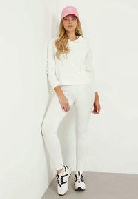 Biela mikina s predným vreckom, zladené biele legíny a čierno-biele tenisky. Rúžová čiapka pridáva farebný akcent.