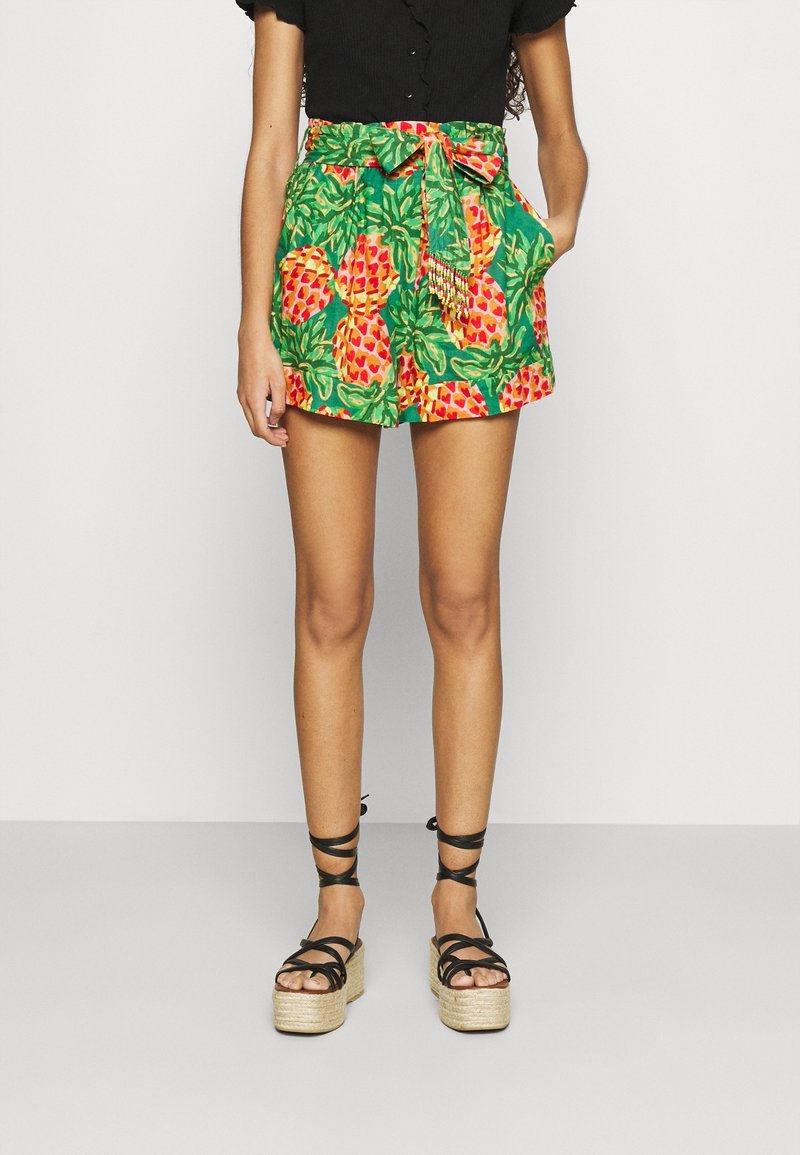 Osoba stoi w czarnej bluzce i kolorowych zielonych szortach w ananasy, dopełnionych czarnymi sandałami na platformie z paskami.