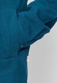 Teal jas met een gestructureerde stof, zijzak en een drukknoopsluiting op de manchetten. Inclusief een klein merklabel.