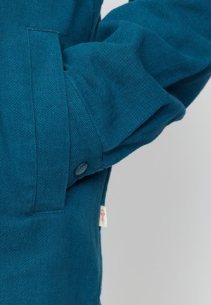 Teal jas met een gestructureerde stof, zijzak en een drukknoopsluiting op de manchetten. Inclusief een klein merklabel.