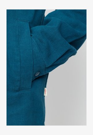 Teal jas met een gestructureerde stof, zijzak en een drukknoopsluiting op de manchetten. Inclusief een klein merklabel.