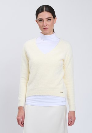 Donna con capelli scuri raccolti in una coda bassa indossa un maglione crema morbido a scollo a V sopra un dolcevita bianco e una gonna chiara su uno sfondo semplice.