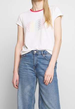 Femme portant un t-shirt blanc avec des lettres multicolores superposées et un jean taille haute bleu, sur un fond clair uni.