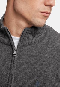 Suéter gris de punto con un alto cuello acanalado y cierre de cremallera. Presenta un logotipo sutil y una tela texturizada en una vista en primer plano.