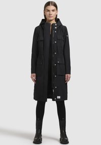 Parka neagră impermeabilă cu glugă, închidere cu capse și fermoar, două buzunare frontale, talie strânsă și design până la genunchi.