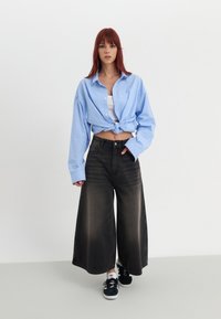 Camisa oversized às riscas azul claro, atada na cintura, combinada com jeans de perna larga pretos. Ténis pretos com detalhes brancos.