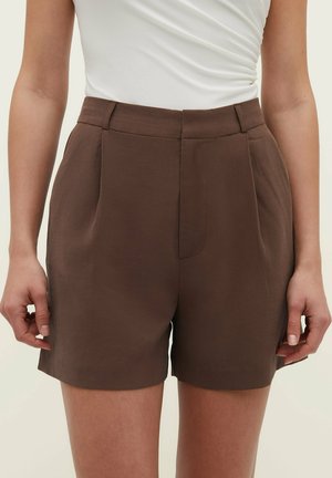 Kvinde iført højttaljede brune skræddersyede shorts med plisseringer og sidelommer, kombineret med en hvid ærmeløs top.