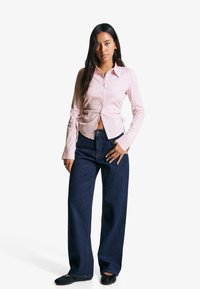 Camicia a maniche lunghe color rosa chiaro con abbottonatura, abbinata a jeans a gamba larga blu navy e scarpe slip-on nere.