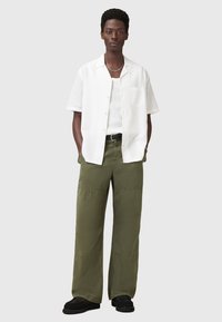 Chemise blanche à manches courtes, pantalons larges vert olive, chaussures noires avec une surface texturée, et un collier chaîne en argent.
