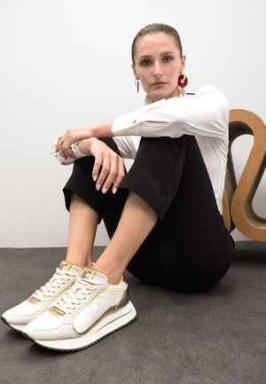 Zapatillas blancas con acentos dorados, con un diseño perforado, cierre de cordones y suela negra. Modelo sentado con las piernas cruzadas, vistiendo una blusa blanca y pantalones negros.