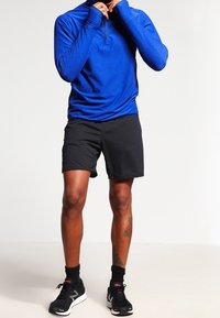 Blå långärmad träningströja med halvdragkedja, ihopparad med svarta sportshorts. Modell som bär svarta sneakers och ankellånga strumpor.