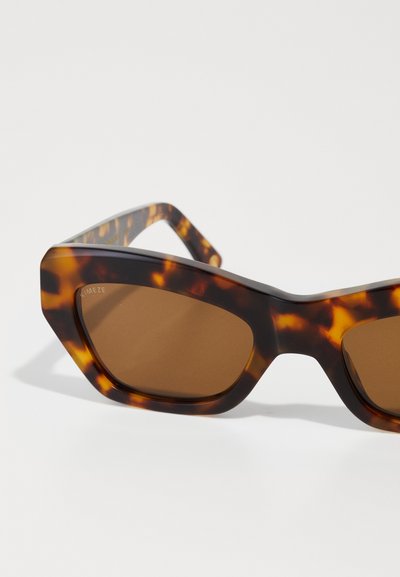 Tortoiseshell sunglasses with a geometric shape, brown lenses, and thick, angular frames. Accents include a patterned texture on the arms.