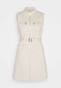 Sleeveless cream dress with a zip front, two zippered chest pockets, a belted waist, and large side pockets. Smooth fabric and tailored fit.