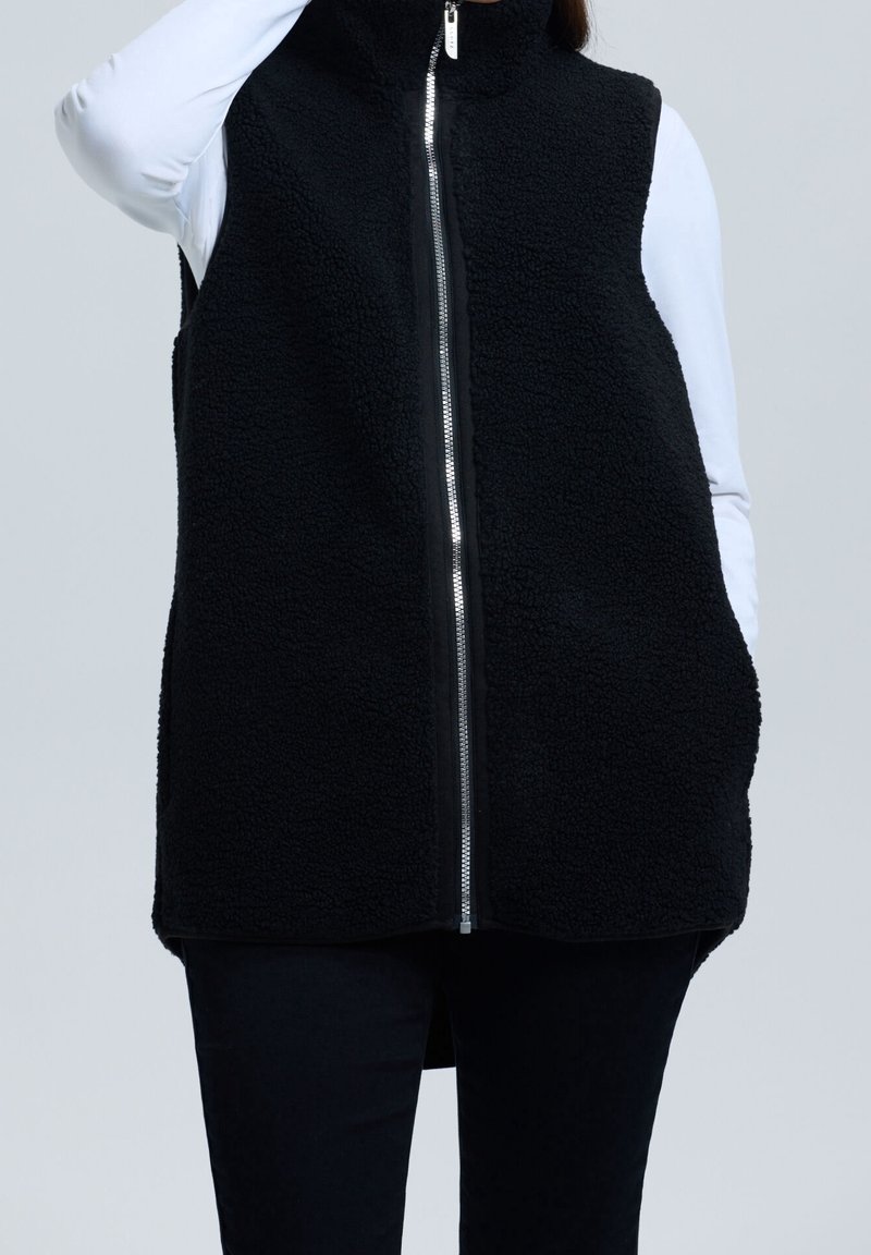 Personne portant un gilet polaire noir sans manches avec une fermeture éclair argentée à l'avant, par-dessus une chemise blanche à manches longues et un pantalon noir.