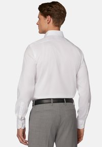 Bela srajca z dolgimi rokavi in zapetimi manšetami, v kombinaciji z sivimi hlačami in črnim pasom, prikazana od zadaj.