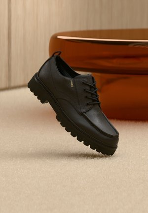 Black leather lace-up shoe with thick rugged sole and gold "HUGO" branding, placed on beige carpet with blurred brown chair in background.