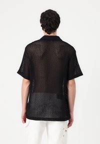 Schwarzes, offen gestricktes Hemd mit Kragen und kurzen Ärmeln, das eine lockere, atmungsaktive Struktur sowie einen etwas längeren Rückensaum aufweist.