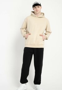 Młody mężczyzna w beżowym hoodie, czarnych spodniach, białych trampkach i czarno-białej czapce, stojący z rękami w kieszeni hoodie.