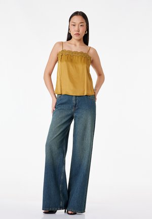 Jeune femme debout, les mains dans les poches, portant un caraco moutarde à dentelle et un jean bleu à jambes larges sur fond blanc.