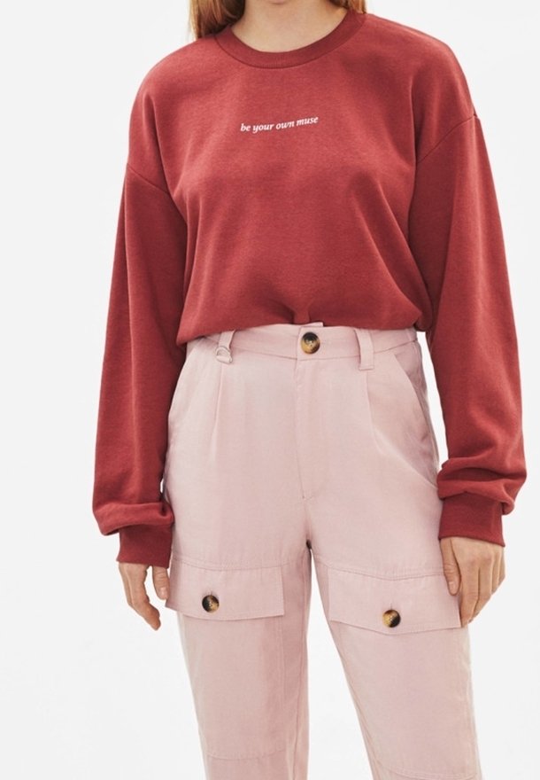 Femme portant un sweat-shirt rouge avec le texte "be your own muse" associé à un pantalon taille haute rose clair avec poches avant boutonnées.