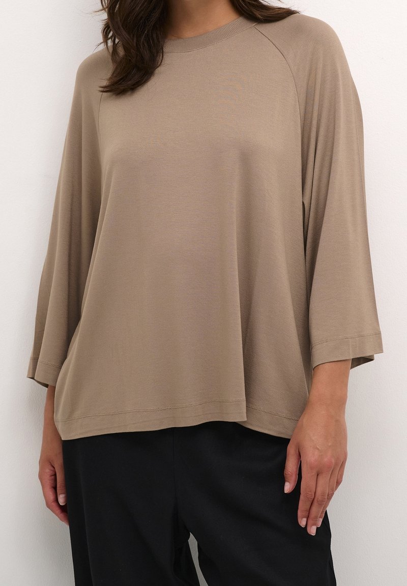 Žena v volném, béžovém topu s dlouhými raglánovými rukávy a černých kalhotách, stojící proti bílé zdi.