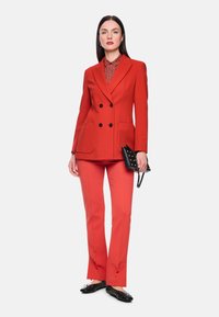 Roter doppelseitiger Anzug mit strukturierte m Stoff, ausgestellte Hose und eine gemusterte Bluse darunter. Accessoires umfassen eine kleine schwarze, mit Nieten besetzte Tasche.