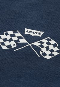 Mørkeblått stoff med et hvitt design av kryssede rutete flagg og Levi's-logoen over, med en jevn tekstur.